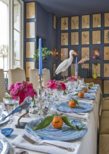 Set table with white table cloth, place settings with blue plates and orange fruit on top.  Table shows taxidermy birds and pink floral arrangements.