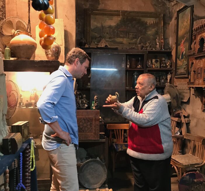 Jamie speaking to man in colorful shop lined with paintings