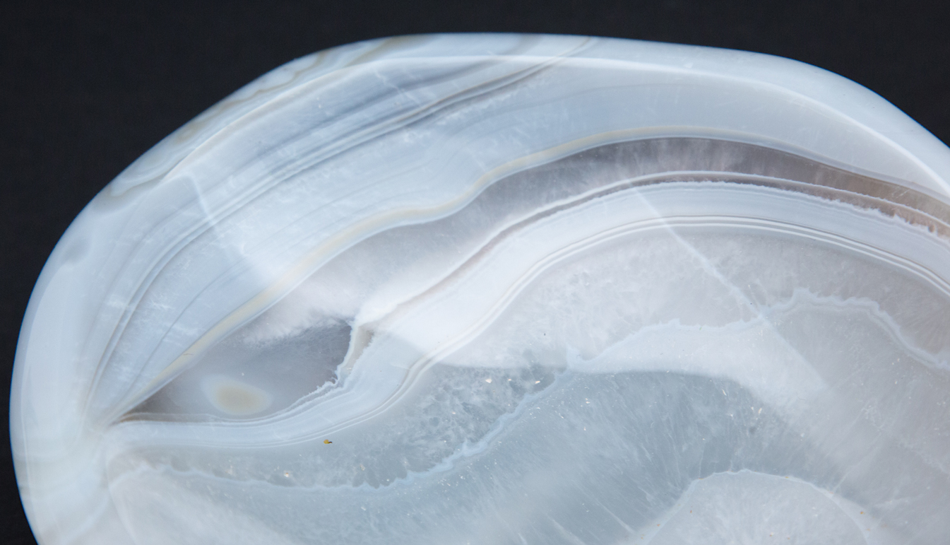 closeup of Agate vide poche with bands of light blue, white, brown, with amorphous shape. Resting on black background