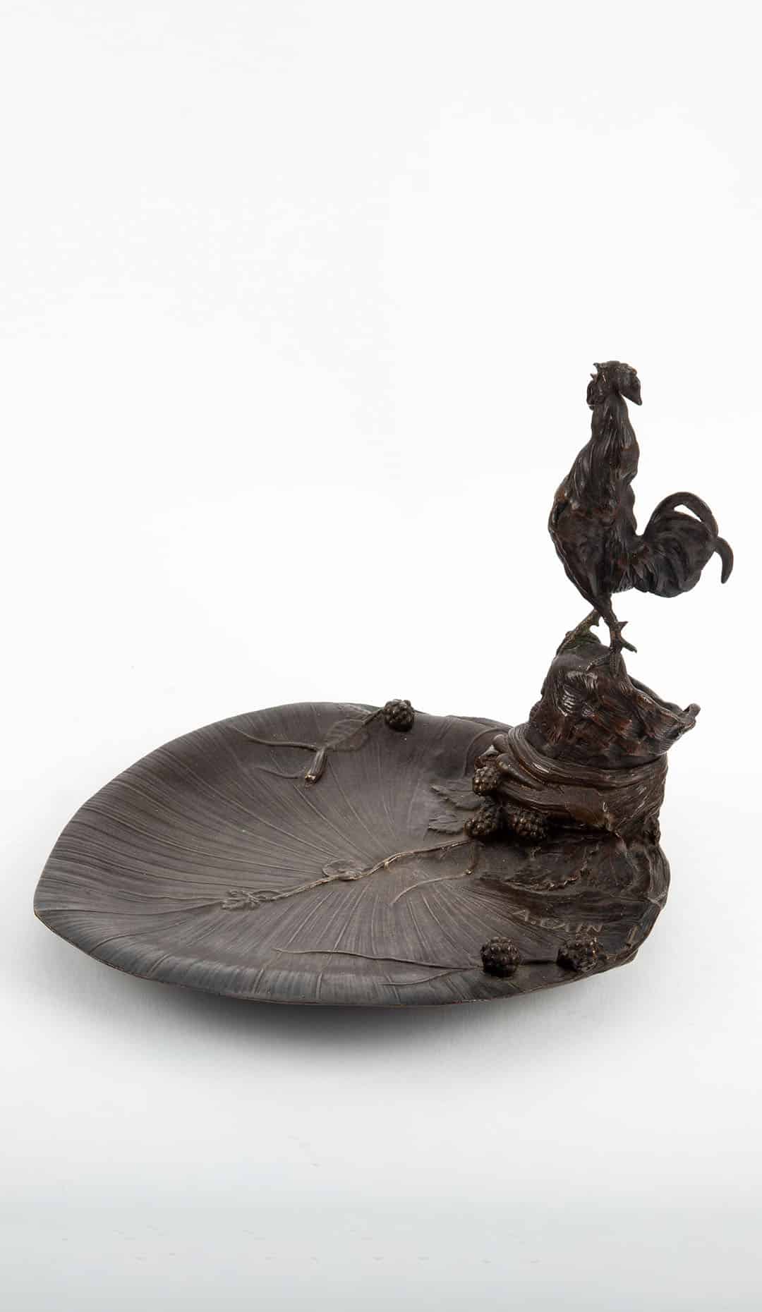 This finely cast 19th-century French bronze vide-poche takes the form of a broad, naturalistically modeled leaf, animated by a sculptural rooster perched upon a rocky outcrop at one end. The surface is richly textured with radiating veins, trailing stems, and small botanical details, demonstrating the high level of craftsmanship associated with French animalier bronzes of the period. The piece is signed “A. Cain”, identifying it as the work of Auguste Cain, one of the most celebrated French animal sculptors of the 19th century. Cain, a student of Alexandre Guionnet and a frequent collaborator of Pierre-Jules Mêne, was renowned for his dynamic and naturalistic depictions of animals, works of which were exhibited at the Paris Salon and widely collected. The rooster—an enduring symbol of France—is rendered with vitality and precision, while the leaf-form tray reflects the period’s fascination with nature-inspired decorative objects. Intended for use as a vide-poche or desk tray, the object successfully balances sculptural presence with practical function. Executed in patinated bronze with a deep, warm surface, this piece exemplifies the refined decorative arts of late 19th-century France and stands as a fine example of Auguste Cain’s animalier output.